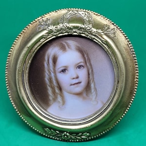 May include: A small, antique portrait of a young girl with blonde hair, set within a round, gold-toned frame. The frame features a decorative laurel wreath at the top and floral accents at the bottom. The portrait is a miniature painting.