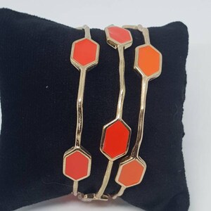 May include: A set of four gold bangles with orange hexagonal accents. The bangles are stacked on top of each other and are displayed on a black surface.