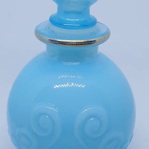 May include: A light blue glass bottle with a round body and a swirl design. The bottle has a gold rim and a light blue stopper with a swirl design.