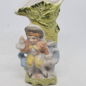 May include: A small ceramic vase shaped like a tree with a boy figurine sitting on a branch. The vase is painted in shades of green, blue, yellow, and pink.
