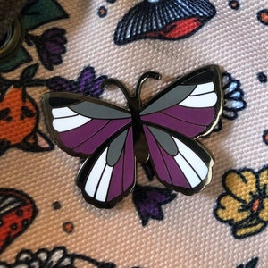 May include: A purple, white, and black butterfly pin on a pink fabric bag with a floral pattern. The bag has a drawstring closure and a brown strap.