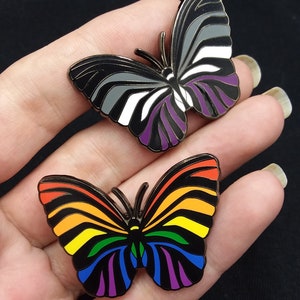 May include: Two enamel pin badges in the shape of butterflies. The first butterfly is black, white, and purple, representing the asexual pride flag. The second butterfly is rainbow colored, representing the LGBTQ+ pride flag.
