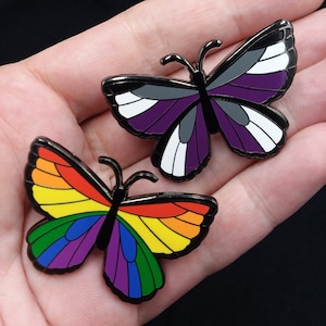 40 mm butterfly pins lined with black nickel. One butterfly has a full rainbow of colors; the other has Ace flag colors: black, gray, white and purple.