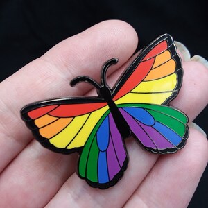 May include: A rainbow-colored butterfly pin with black outlines. The butterfly has a black body and wings with a rainbow gradient.