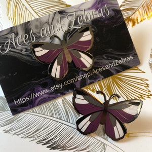 May include: Asexual pride flag butterfly pin with purple, black, and white stripes. The pin is on a white background with gold and silver feathers.