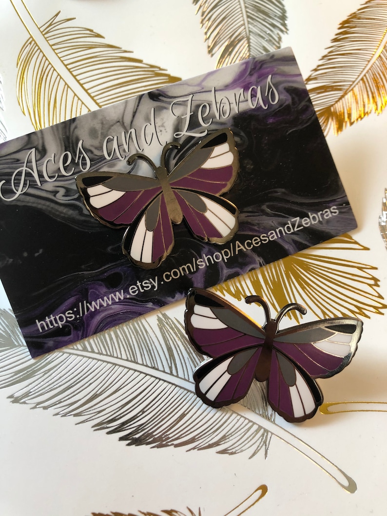 May include: Asexual pride flag butterfly pin with purple, black, and white stripes. The pin is on a white background with gold and silver feathers.