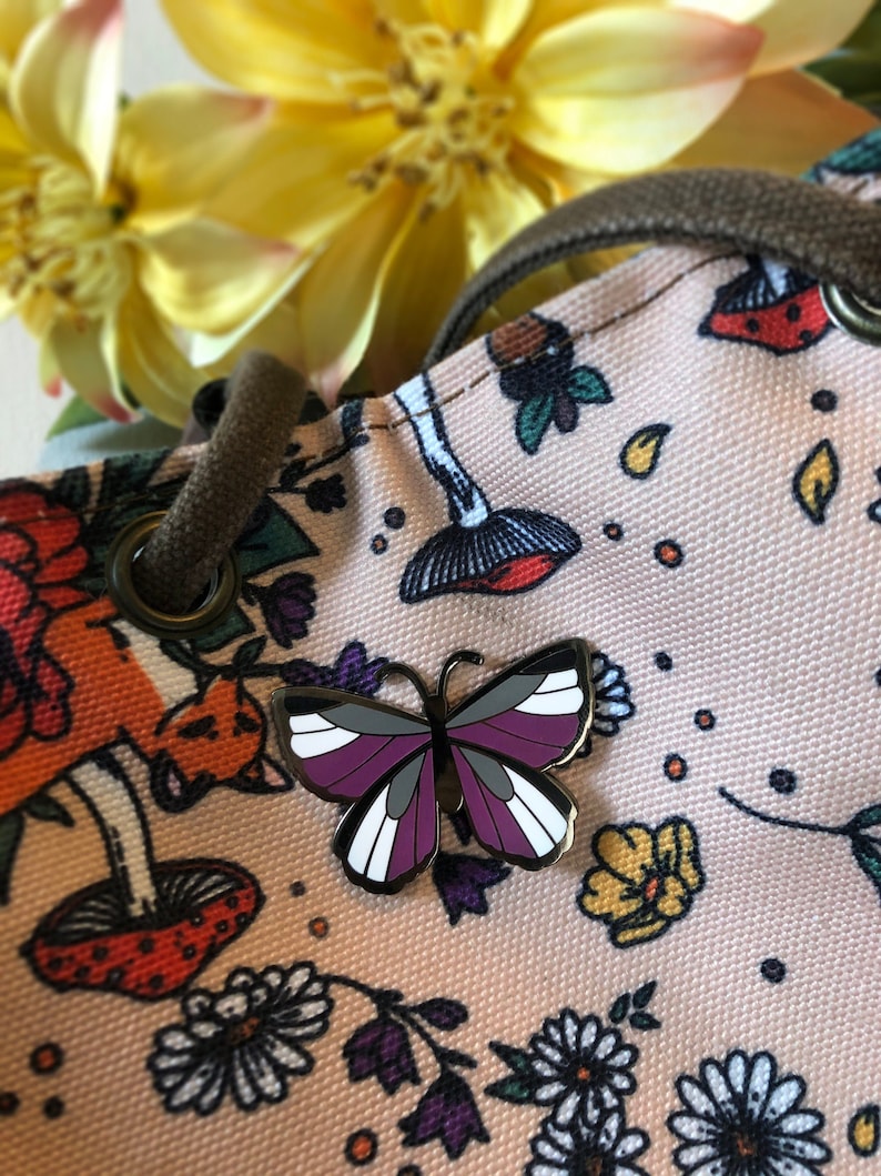May include: A purple, white, and black butterfly pin on a pink fabric bag with a floral pattern. The bag has a drawstring closure and a brown strap.