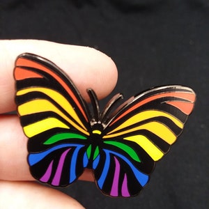 May include: A rainbow-colored butterfly pin with black stripes and a black outline. The butterfly is made of metal and has a shiny finish.