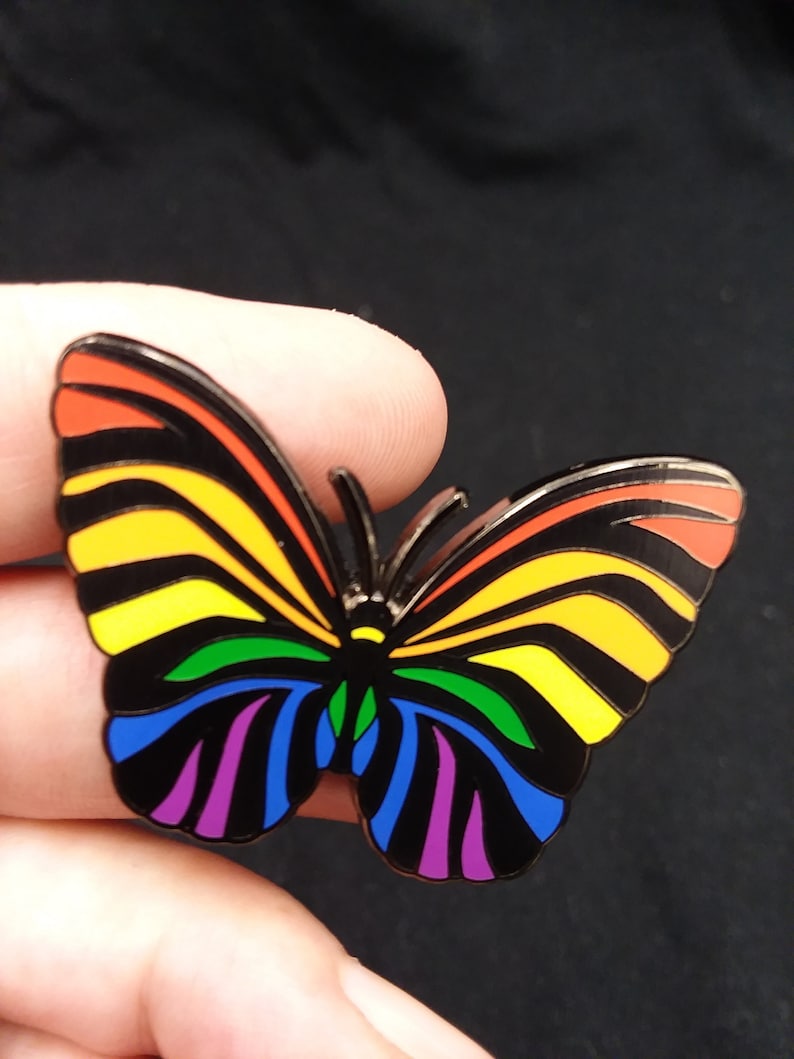 May include: A rainbow-colored butterfly pin with black stripes and a black outline. The butterfly is made of metal and has a shiny finish.