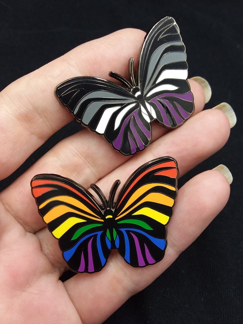 May include: Two enamel pin badges in the shape of butterflies. The first butterfly is black, white, and purple, representing the asexual pride flag. The second butterfly is rainbow colored, representing the LGBTQ+ pride flag.