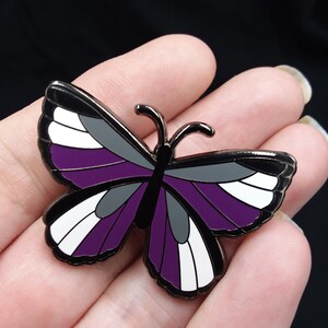 May include: A black metal butterfly pin with purple, white, and gray stripes. The pin is a symbol of asexuality.