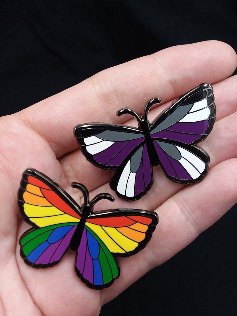 40 mm butterfly pins lined with black nickel. One butterfly has a full rainbow of colors; the other has Ace flag colors: black, gray, white and purple.