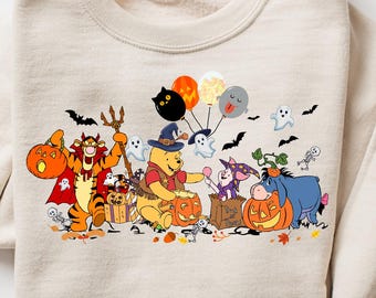 May include: A cream-coloured sweatshirt with a Halloween-themed cartoon graphic. The design features characters like Winnie the Pooh, Tigger, and Eeyore in costumes, with pumpkins, bats, and balloons. The sweatshirt is folded with rolled-up sleeves.