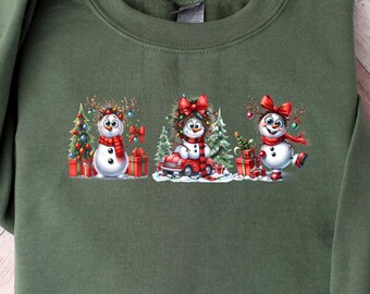 May include: A green sweatshirt with a Christmas-themed design featuring three snowmen wearing red hats and scarves. The snowmen are surrounded by Christmas trees, presents, and a sled.