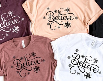 May include: Five t-shirts with the word "Believe" in a cursive font with snowflakes around the word. The shirts are in various colors including maroon, beige, black, and white.