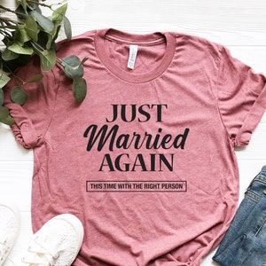 May include: A pink t-shirt with the text "Just Married Again" and the smaller text "This time with the right person" printed on it. The shirt is laid flat on a white surface with a pair of white sneakers and a pair of blue jeans.