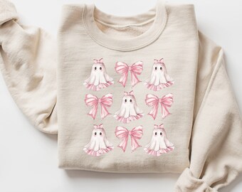 May include: A beige sweatshirt with a pattern of white ghosts wearing pink bows. The ghosts are arranged in a grid pattern.