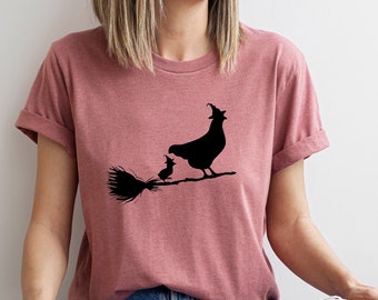 May include: A woman wearing a mauve t-shirt with a black silhouette of a chicken wearing a witch's hat and holding a broom.