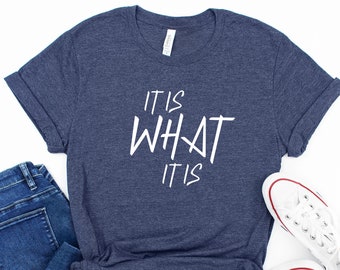 May include: A navy blue t-shirt with the text "IT IS WHAT IT IS" in white lettering. The shirt is paired with blue jeans and white sneakers.