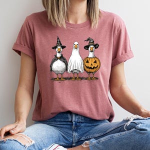 May include: A dusty rose t-shirt featuring a Halloween-themed graphic. The graphic depicts three geese dressed in costumes: a witch, a ghost, and a jack-o'-lantern. The geese are standing side-by-side, with their costumes detailed in black and orange.
