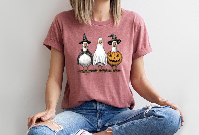 May include: A dusty rose t-shirt featuring a Halloween-themed graphic. The graphic depicts three geese dressed in costumes: a witch, a ghost, and a jack-o'-lantern. The geese are standing side-by-side, with their costumes detailed in black and orange.