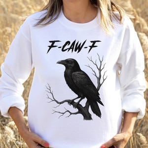 May include: White long-sleeve sweatshirt featuring a black raven perched on a branch. The text "F-CAW-F" is printed above the raven. The raven and branch design is in the center of the sweatshirt.