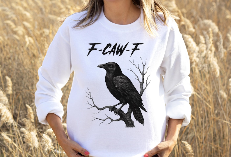 May include: White long-sleeve sweatshirt featuring a black raven perched on a branch. The text "F-CAW-F" is printed above the raven. The raven and branch design is in the center of the sweatshirt.