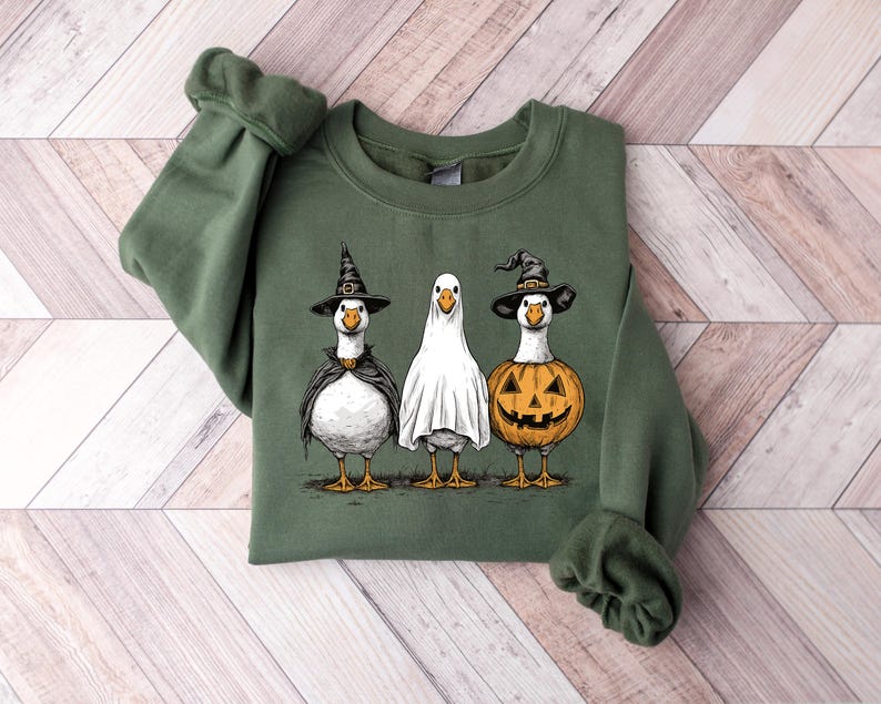 May include: Green sweatshirt featuring three geese dressed in Halloween costumes. One goose wears a witch hat and cape, another is a ghost, and the third is a jack-o'-lantern. The design is a whimsical illustration for the spooky season.