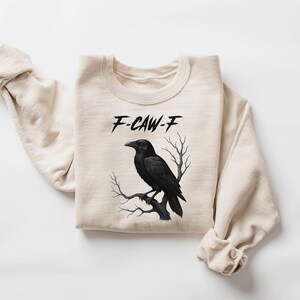 May include: A cream-colored sweatshirt featuring a black raven perched on a branch. The text "F-CAW-F" is above the raven. The design is centered on the front of the folded sweatshirt, with the sleeves casually draped.