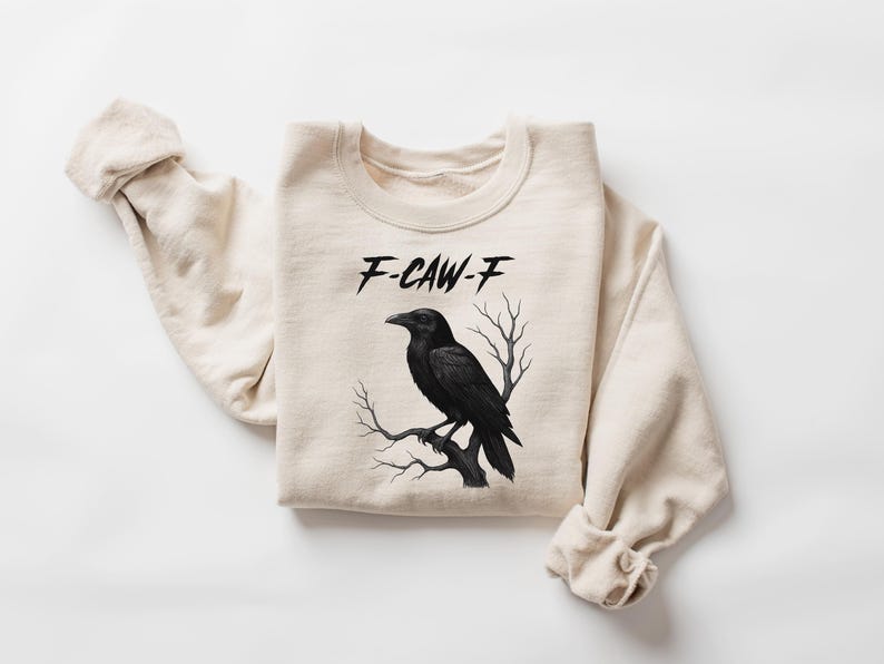 May include: A cream-colored sweatshirt featuring a black raven perched on a branch. The text "F-CAW-F" is above the raven. The design is centered on the front of the folded sweatshirt, with the sleeves casually draped.