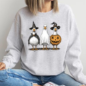 May include: A light gray sweatshirt featuring a Halloween-themed graphic. The graphic depicts three geese dressed in costumes: a witch, a ghost, and a jack-o'-lantern. The geese are standing on a patch of grass.