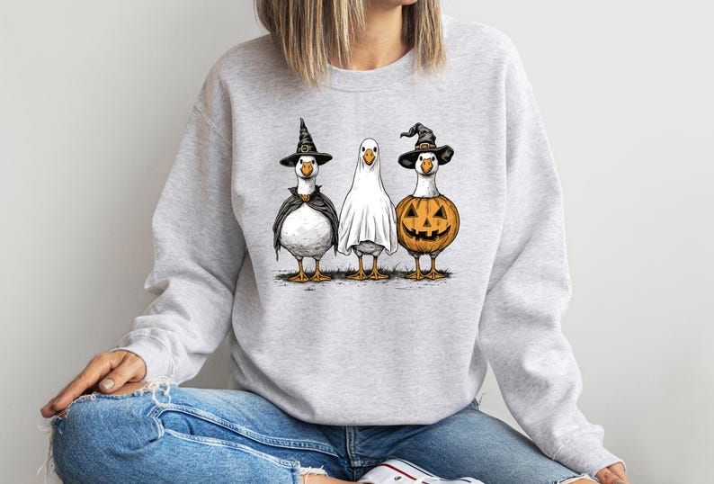 May include: A light gray sweatshirt featuring a Halloween-themed graphic. The graphic depicts three geese dressed in costumes: a witch, a ghost, and a jack-o'-lantern. The geese are standing on a patch of grass.