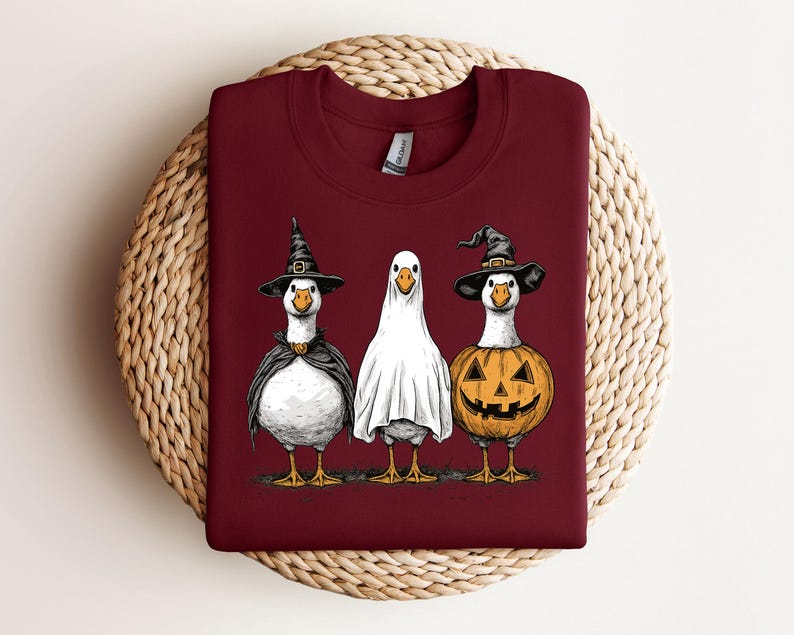 May include: A maroon sweatshirt featuring a Halloween-themed graphic of three geese. One goose is dressed as a witch, another as a ghost, and the third as a jack-o'-lantern. The geese are standing on a patch of grass, with a woven background.
