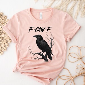 May include: A light pink t-shirt featuring a black raven perched on a branch. The text "F-CAW-F" is above the raven. The shirt has rolled-up sleeves and a round neckline.