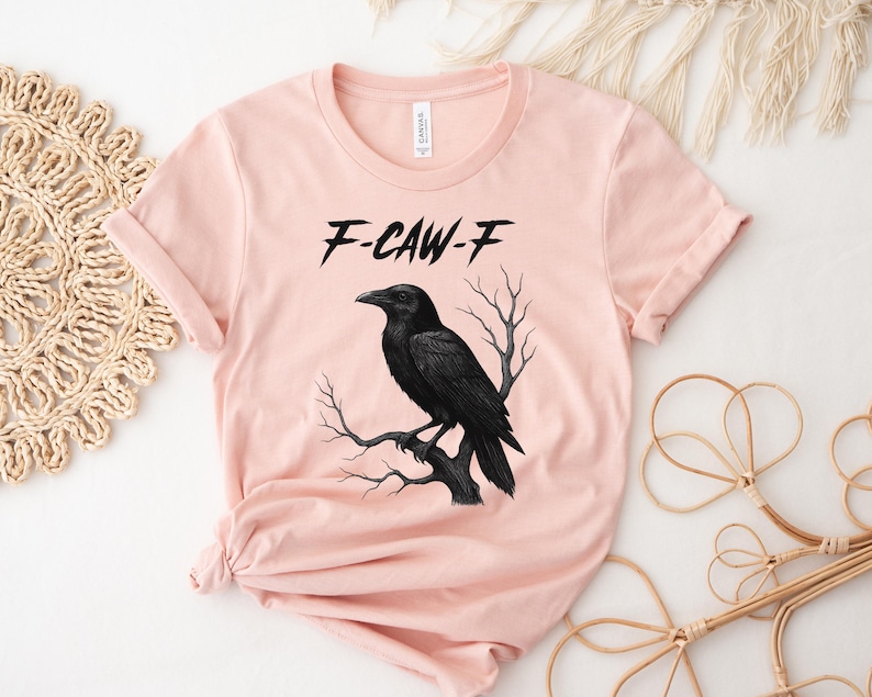 May include: A light pink t-shirt featuring a black raven perched on a branch. The text "F-CAW-F" is above the raven. The shirt has rolled-up sleeves and a round neckline.