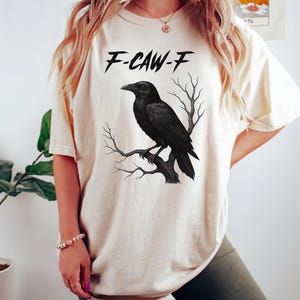 May include: Cream-colored t-shirt featuring a black raven perched on a branch, with the text "F-CAW-F" above the bird. The raven is detailed, and the shirt has a relaxed fit.