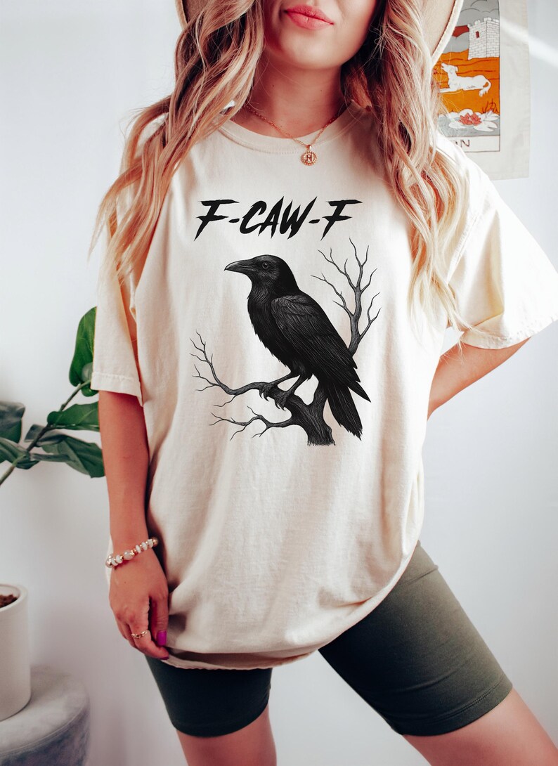 May include: Cream-colored t-shirt featuring a black raven perched on a branch, with the text "F-CAW-F" above the bird. The raven is detailed, and the shirt has a relaxed fit.