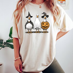 May include: Beige t-shirt featuring three cartoon geese dressed for Halloween. One goose wears a witch hat and cape, another is a ghost, and the third is a jack-o'-lantern. The design is a whimsical illustration.