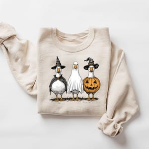 May include: Beige sweatshirt featuring three geese dressed for Halloween. One goose wears a witch hat and cape, another is a ghost, and the third is a jack-o'-lantern. The graphic is in black, orange, and white.