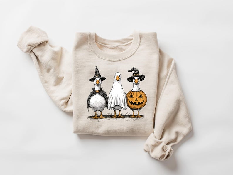May include: Beige sweatshirt featuring three geese dressed for Halloween. One goose wears a witch hat and cape, another is a ghost, and the third is a jack-o'-lantern. The graphic is in black, orange, and white.