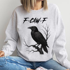 May include: Light gray sweatshirt with a black raven perched on a branch. The text "F-CAW-F" is above the raven. The sweatshirt is worn with blue jeans.