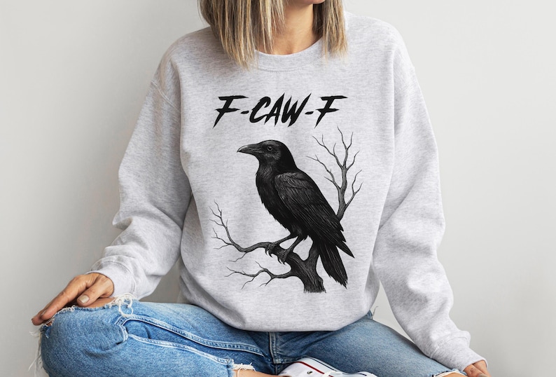 May include: Light gray sweatshirt with a black raven perched on a branch. The text "F-CAW-F" is above the raven. The sweatshirt is worn with blue jeans.
