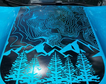 May include: A blue Jeep with a black decal on the hood. The decal features a mountain range with trees and a topographic map design. The Jeep logo is visible on the front of the vehicle.