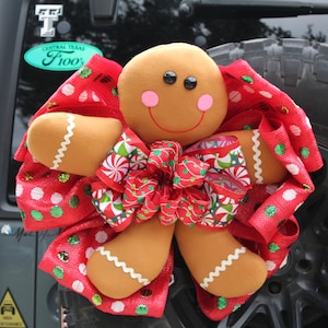 May include: A gingerbread man Christmas wreath with a red, white, and green bow. The wreath is attached to the back of a black vehicle.