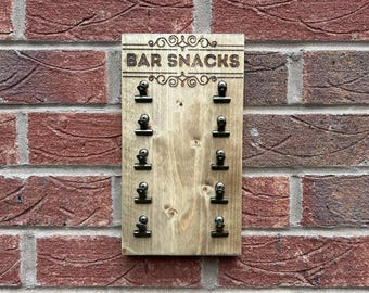 May include: A rectangular light brown wooden board featuring the engraved text BAR SNACKS at the top with decorative scrollwork. Ten small black metal clips are arranged in two vertical columns of five, each secured with a silver screw.