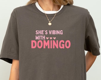 May include: A dark gray t-shirt with the text "SHE'S VIBING WITH DOMINGO" in pink. The shirt has a white collar and a relaxed fit. The text is in a playful font with three pink hearts above the word "DOMINGO".