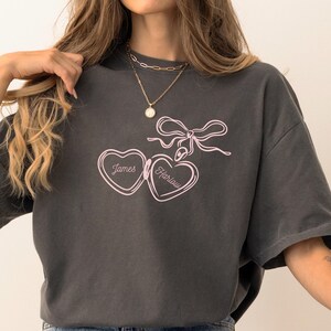 May include: An oversized charcoal grey t-shirt featuring a pink outline of a heart-shaped locket with the names "James" and "Harlow" inscribed on it.