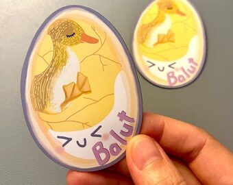 May include: Two stickers featuring a cartoon duckling hatching from an egg. The duckling is yellow and brown, with closed eyes and orange beak. The egg is yellow with cracks, and the word "Balut" is written in purple.