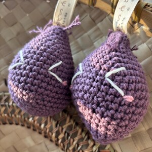 May include: Two handmade, purple crochet ornaments with white and pink accents. The ornaments are teardrop-shaped and have a textured appearance. They hang from a woven basket with a ribbon that has text on it.