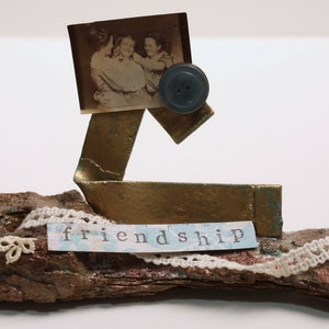 May include: A small, rustic wooden sculpture with a gold metal piece and a vintage photograph of three people. The sculpture is decorated with white lace and the word "friendship" is written on a piece of paper.